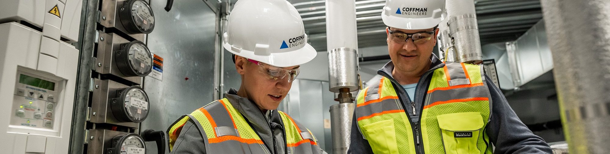 Two Coffman engineers wearing vests and hard hats look over blueprints on a job site.