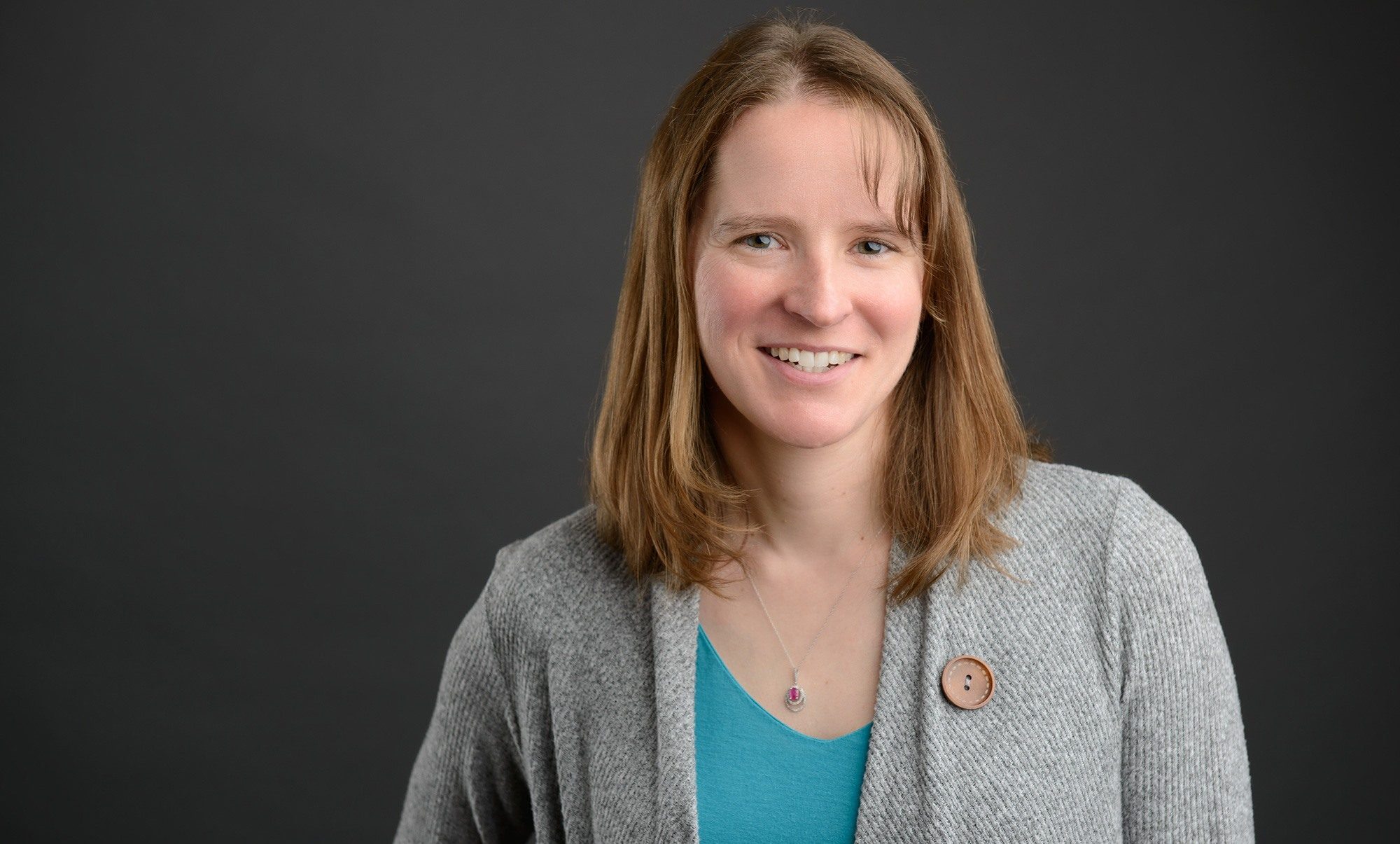 Corrosion Control Engineering Principal Cynthia smiles in front of a black background wearing a blue shirt and grey sweater.
